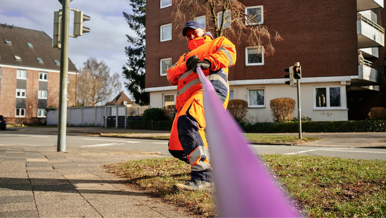 Arbeiter in orangefarbener Schutzkleidung verlegt ein lila Glasfaserkabel an einer Straßenecke.